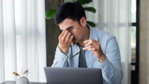 Man pinching tired eyes with glasses off while working at laptop on desk