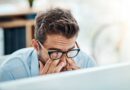 Man with glasses tiredly rubbing eyes while working on a laptop PC
