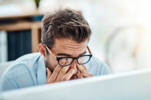 Man with glasses tiredly rubbing eyes while working on a laptop PC