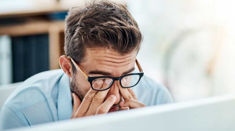 Man with glasses tiredly rubbing eyes while working on a laptop PC